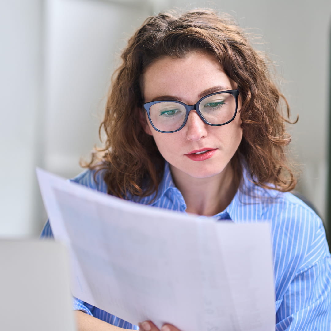 Woman with glasses reading papers at a desk, wearing a blue striped shirt, focused and seated in a bright room.