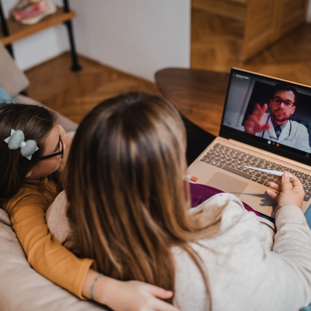 Mother and daughter sitting on a couch, having a video call with a doctor on a laptop.