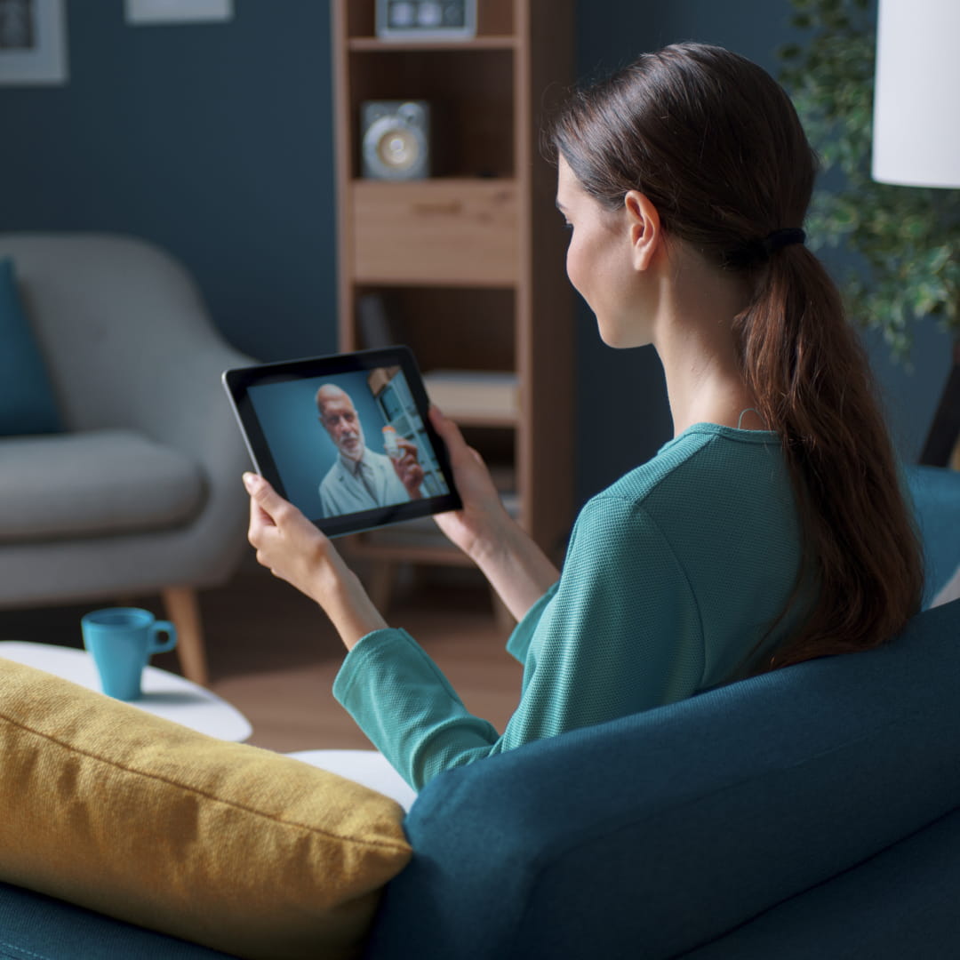 Woman on blue sofa video chatting with a bearded man on a tablet, yellow pillow, cup, and modern living room setting.