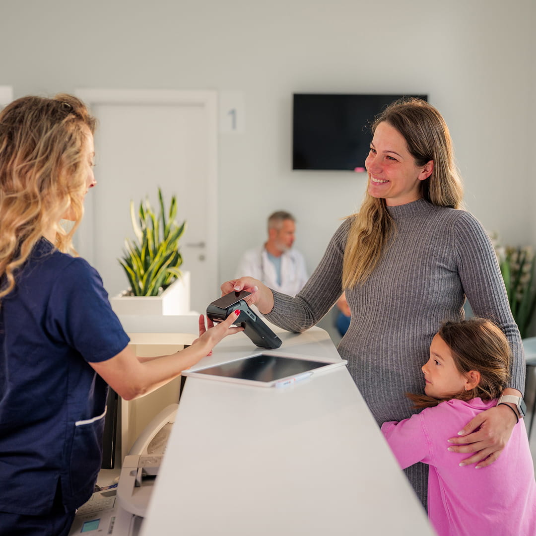 Woman with child at reception desk hands bank card to nurse, plants in background.