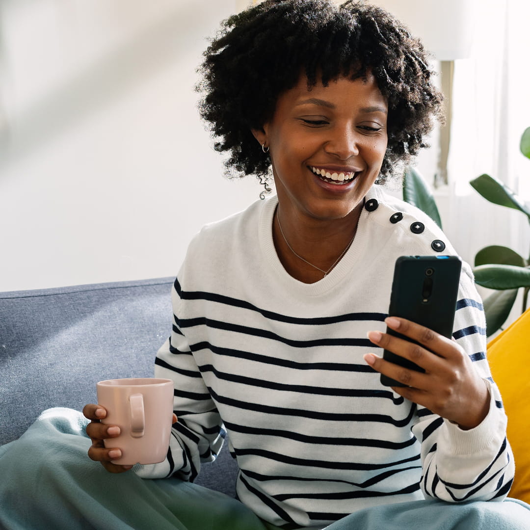 A woman with curly hair smiles at her phone while holding a pink mug, sitting on a sofa with a striped sweater.