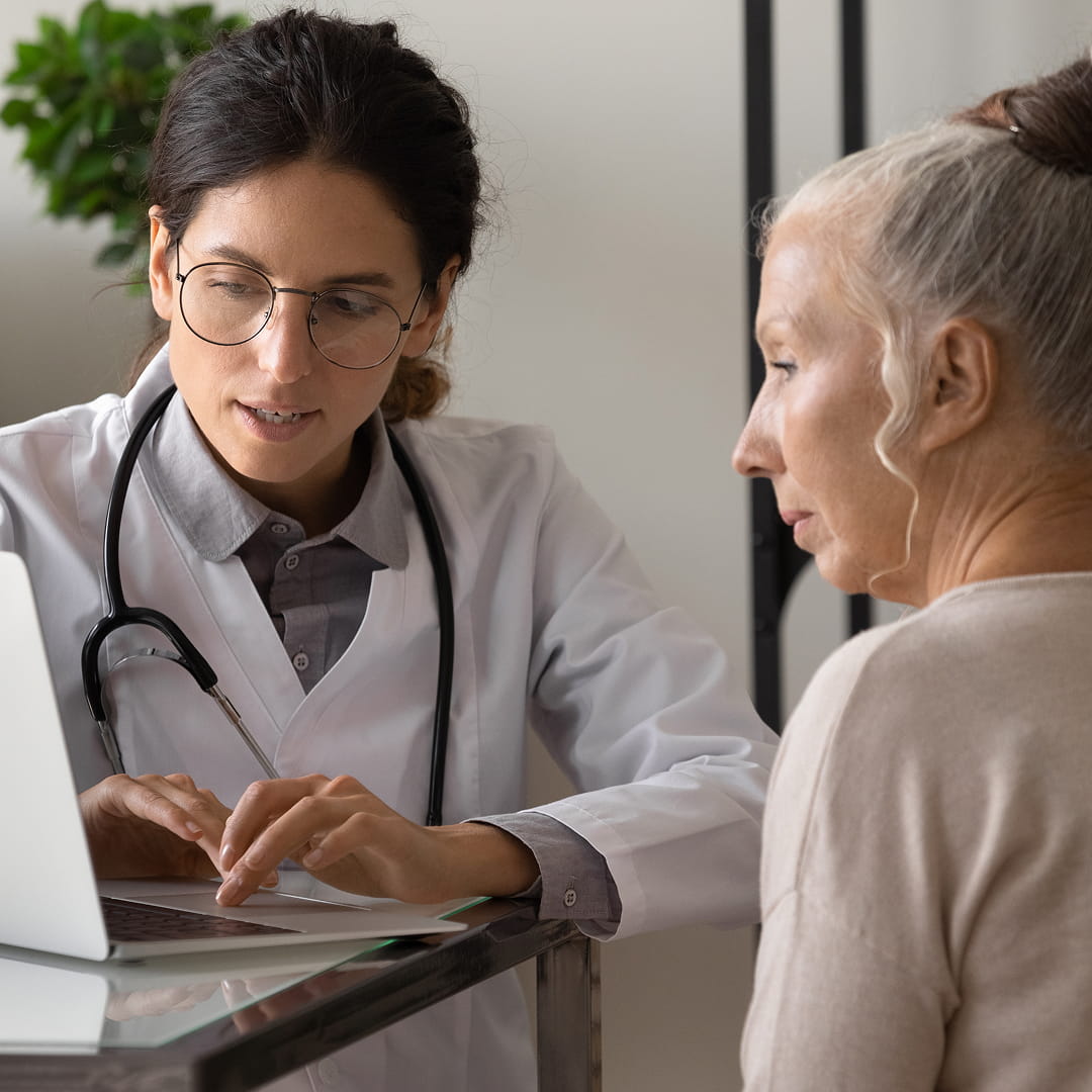 Doctor in white coat discusses information with an older woman at a desk, using a laptop.