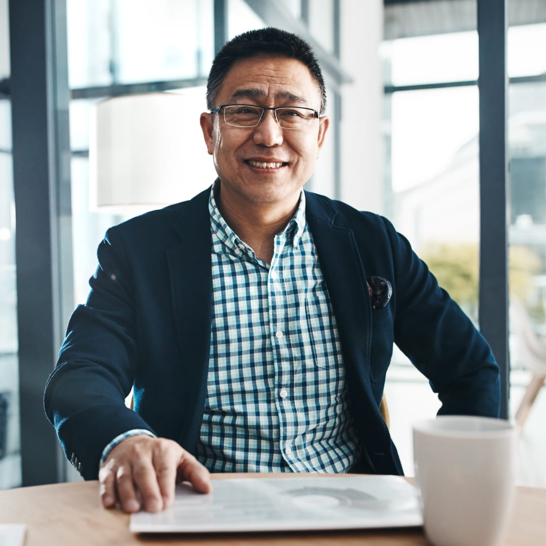Man in a checkered shirt and blazer smiling, sitting at a table with a cup and an open laptop, bright window behind.