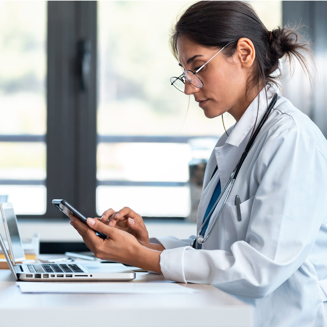 Doctor using a smartphone at a desk with a laptop, wearing glasses and a stethoscope in a bright office.