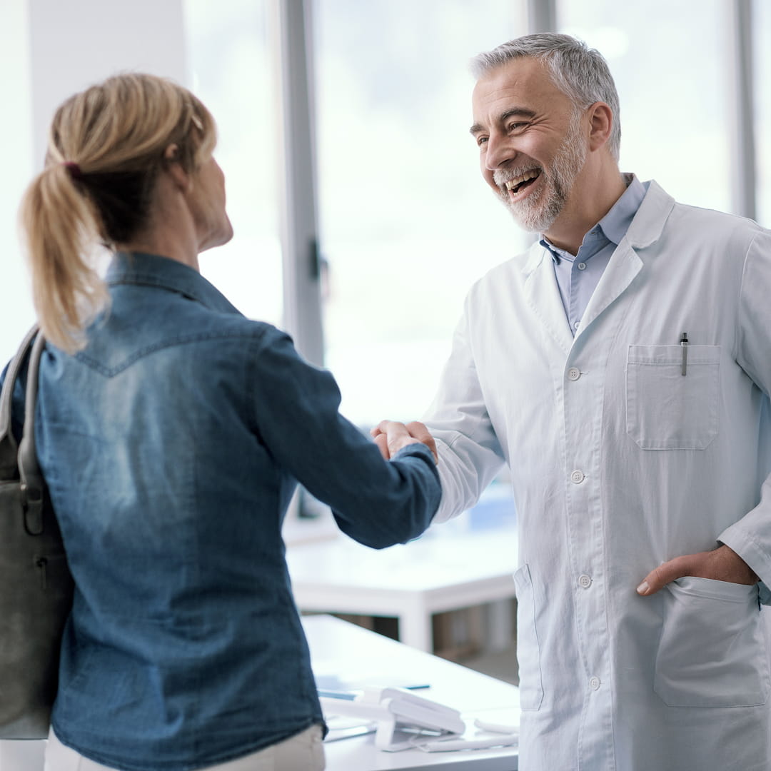A doctor in a white coat smiles while shaking hands with a person wearing a denim jacket in a bright office.