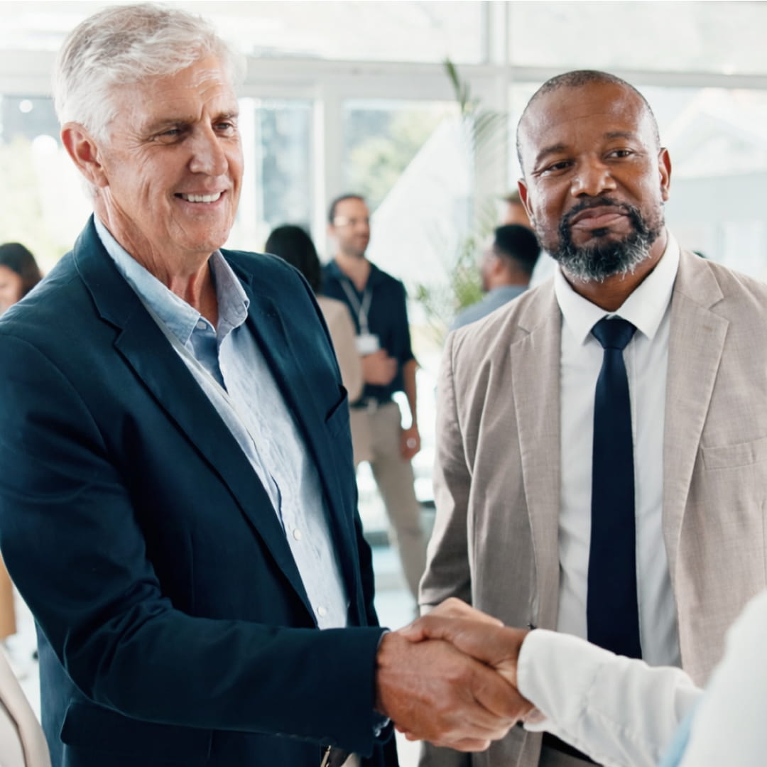 Two men in suits shaking hands while smiling in a bright office environment; people standing in the background.