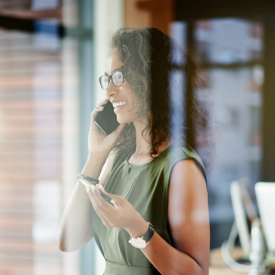 Woman in green dress smiling and talking on phone indoors, holding a pen. Glass reflection partially visible in foreground.