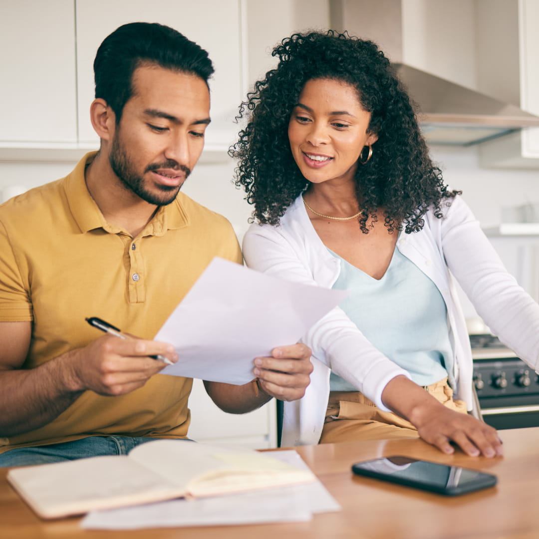 Man and woman sitting at kitchen table, discussing a document with open book and smartphone nearby.