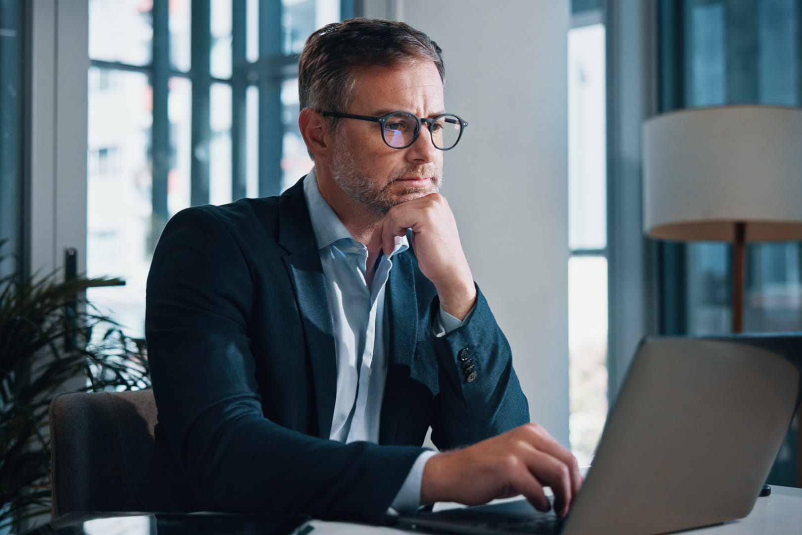 Man in a suit wearing glasses works on a laptop in a modern office, resting his chin on his hand thoughtfully.