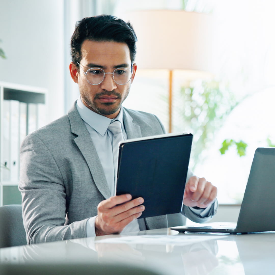 Man in suit and glasses looks at a tablet in a bright, modern office with a laptop on the table.