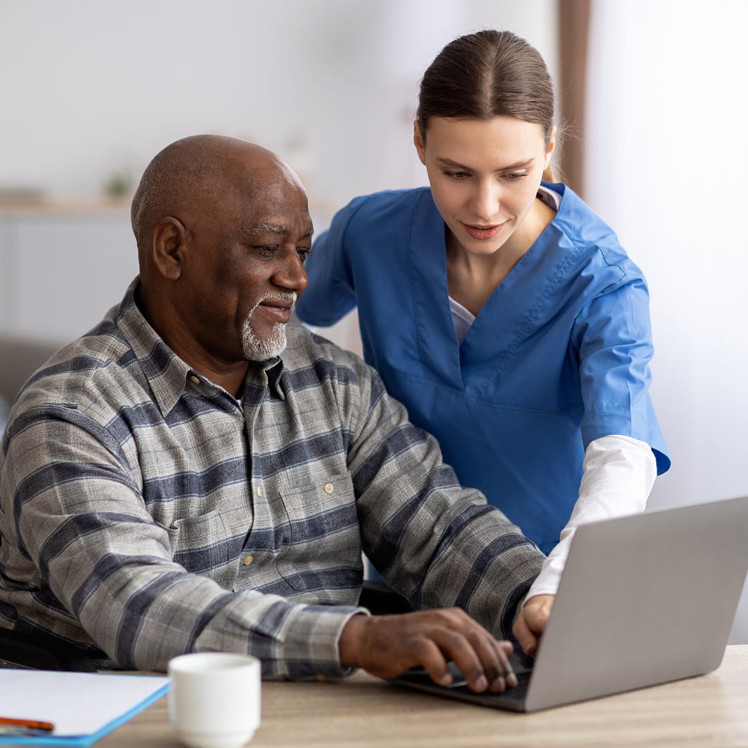 A caregiver in blue scrubs assists an elderly man using a laptop at a table, with a coffee cup nearby.