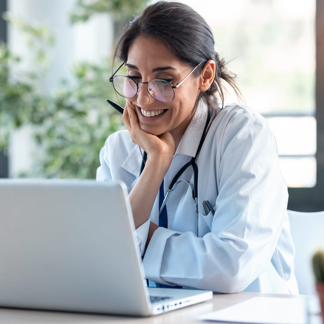 Doctor smiling while looking at a laptop, wearing a white coat and stethoscope, seated at a desk in a bright office.