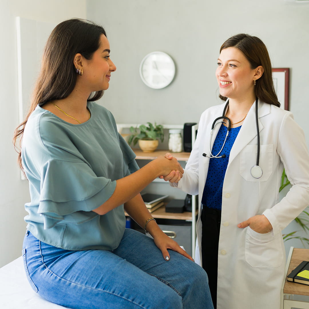 Doctor with stethoscope shakes hands with woman sitting on exam table in medical office.