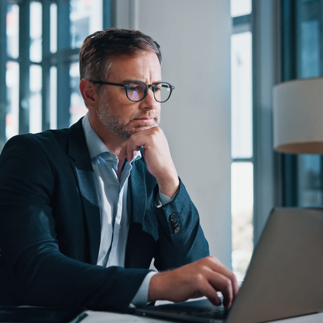 Man with glasses and beard working on a laptop in an office, wearing a suit, hand on chin, looking at screen thoughtfully.