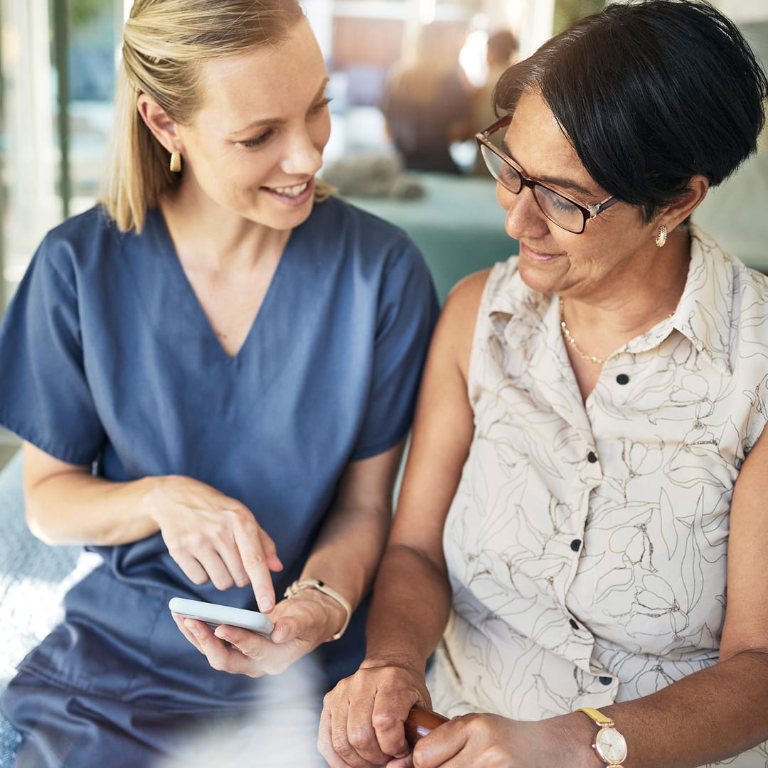 Healthcare worker showing an elderly woman something on a smartphone; they both smile and are seated indoors.