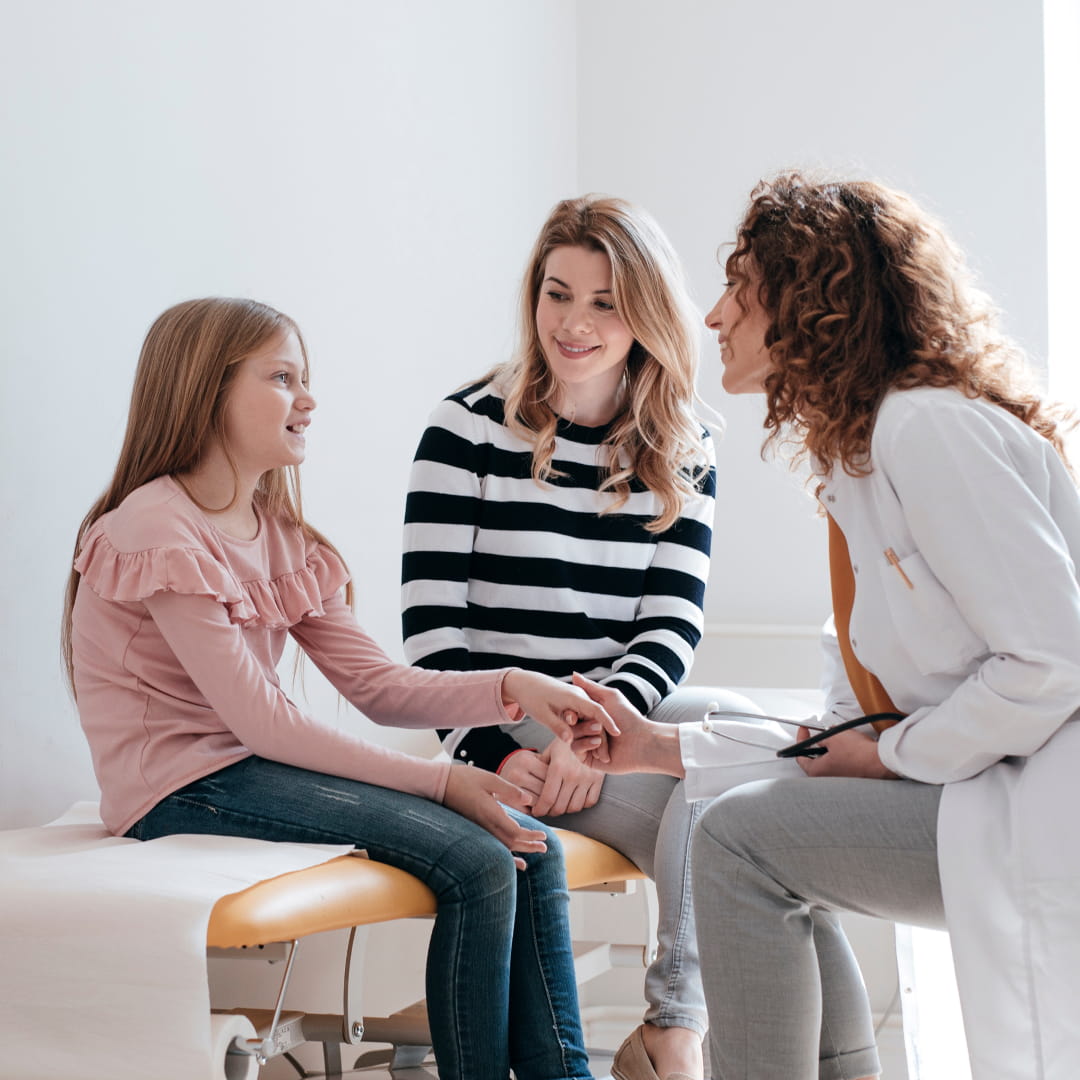 Doctor talking kindly to a young girl sitting with her mother, in a bright medical office.