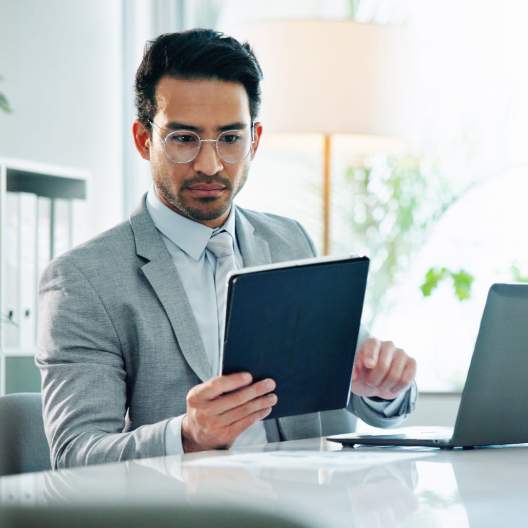 Professional man in gray suit reading a tablet at an office desk with a laptop and soft lighting in the background.