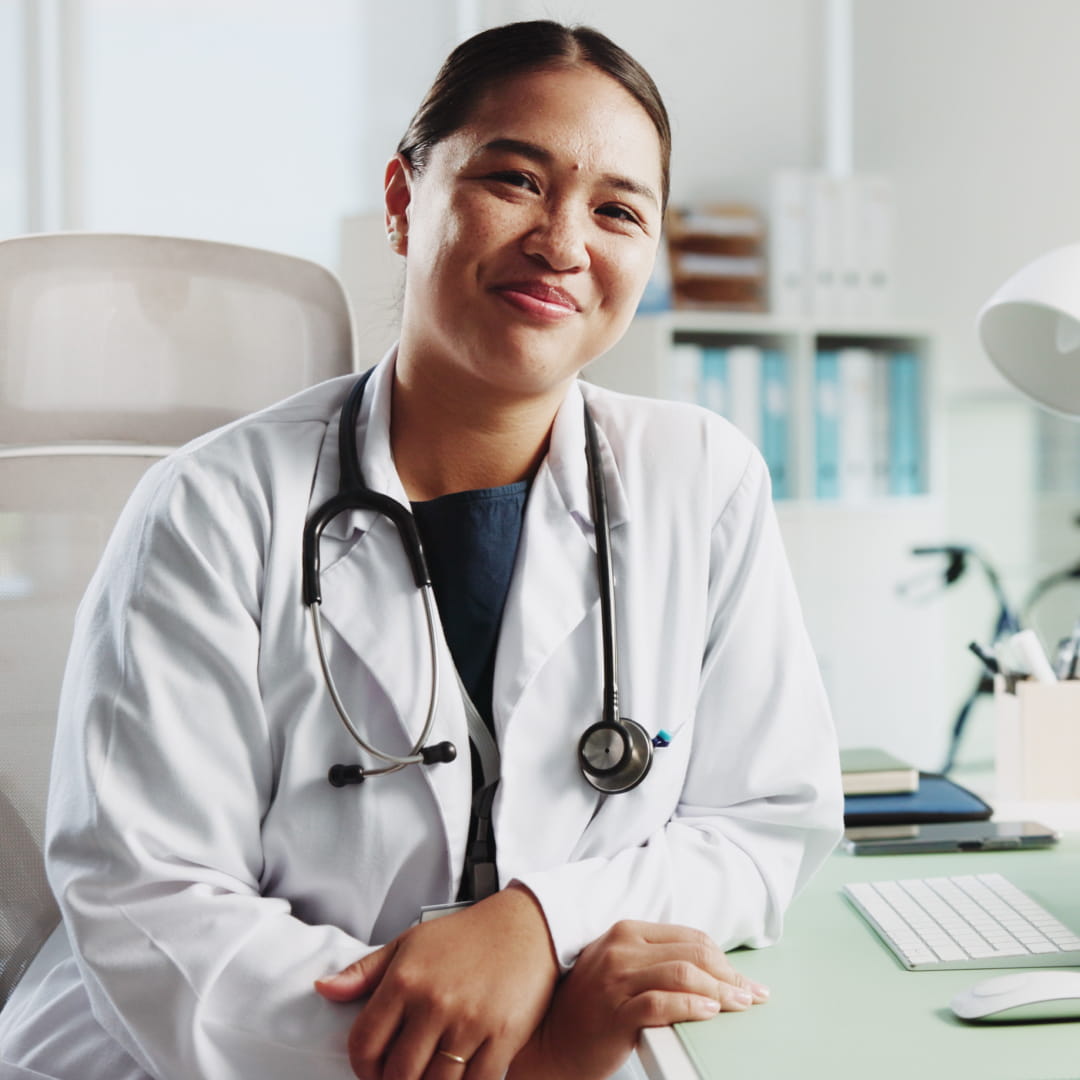 Doctor sitting at a desk with a stethoscope around her neck, smiling in a brightly lit office environment.