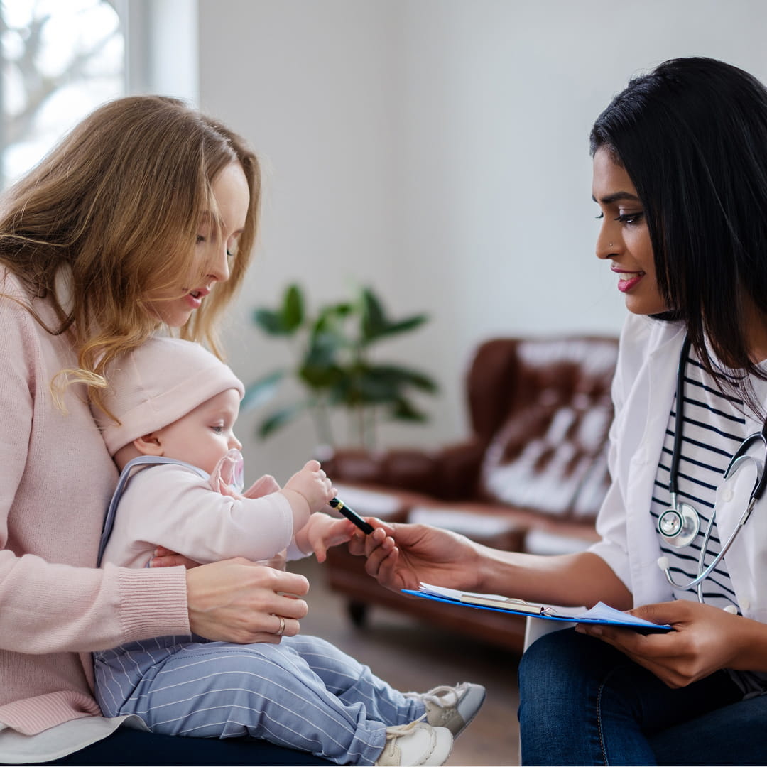 Doctor with stethoscope handing clipboard to woman holding baby in living room setting.