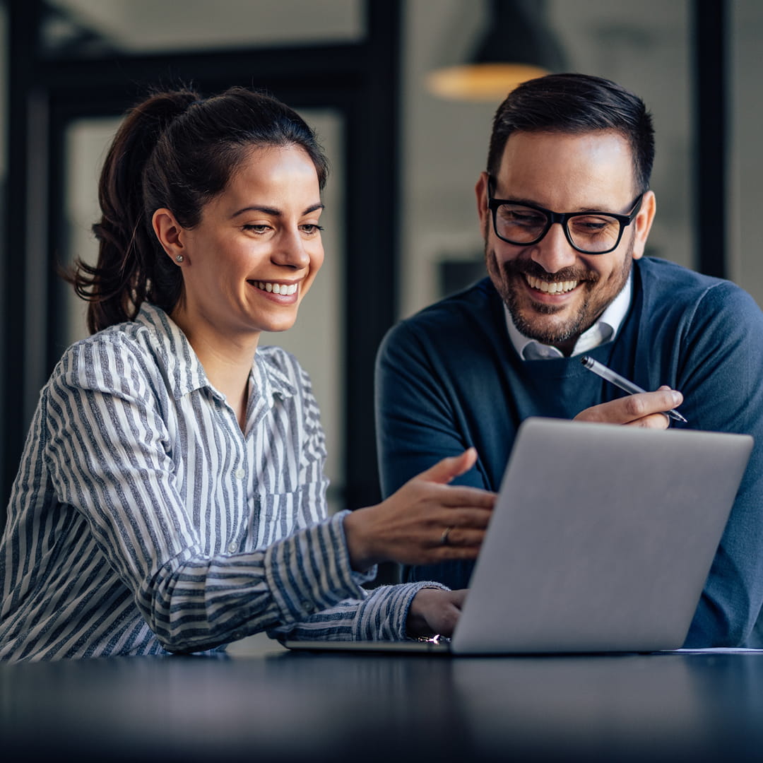 Two colleagues smiling and discussing work on a laptop in a modern office setting.