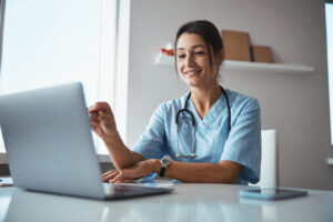 Cheerful young woman physician sitting at the table and smiling while pointing pen at notebook display