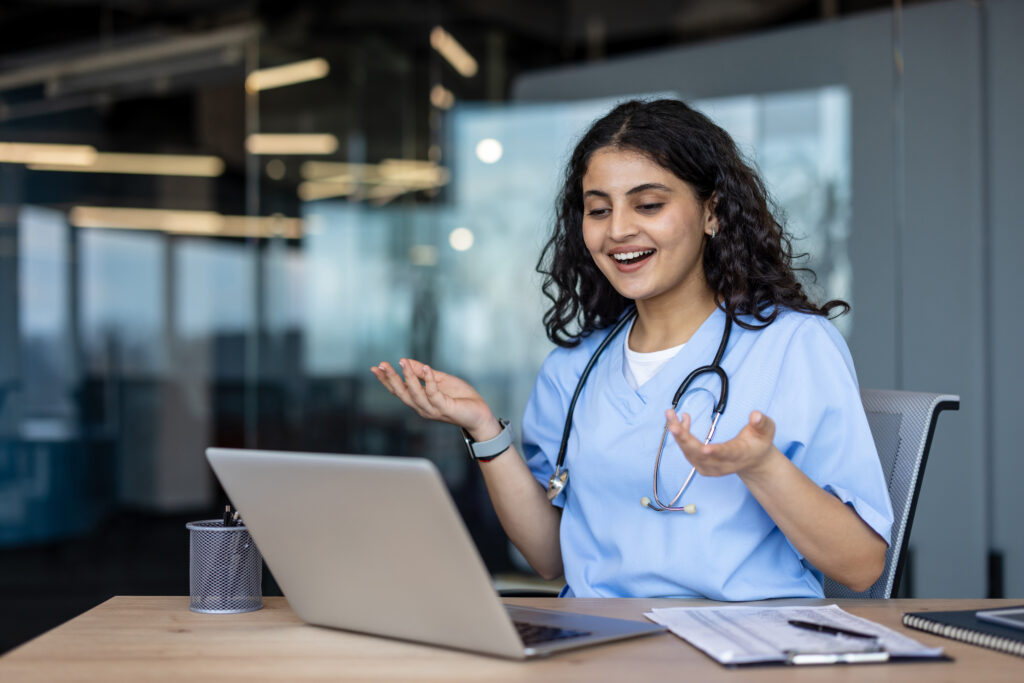 Young indian female doctor in scrubs and stethoscope smiling and gesturing while having a video call on her laptop, providing an online medical consultation from a modern office