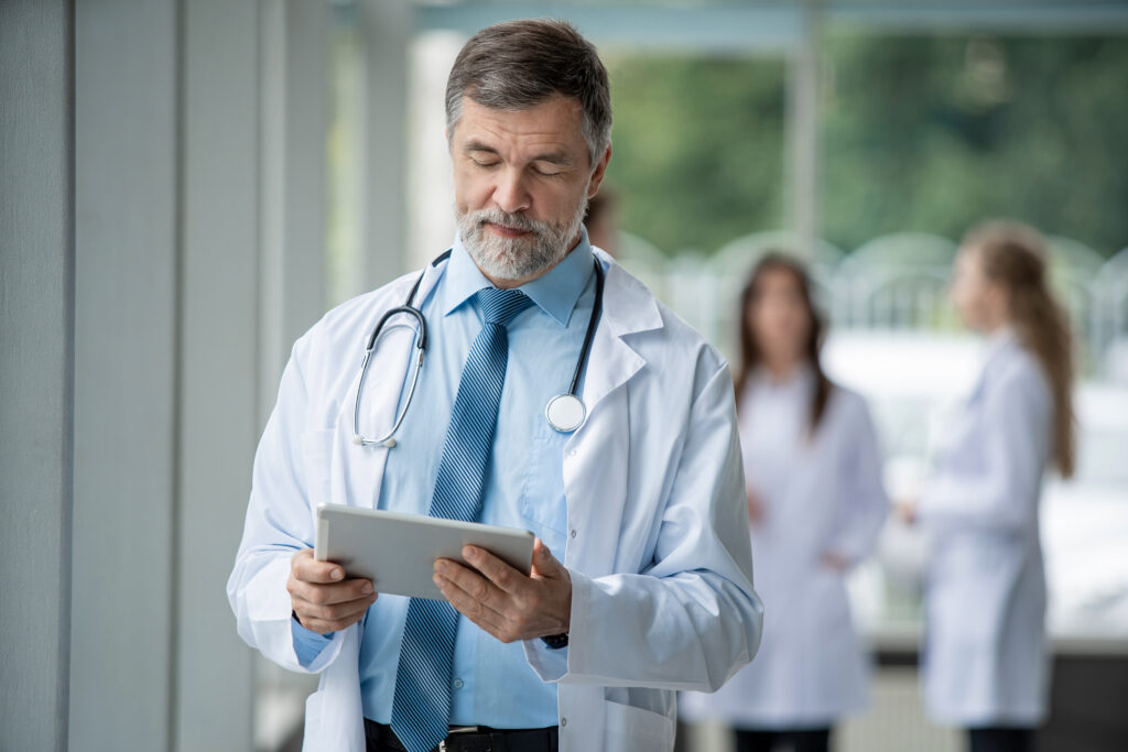 Confident smiling friendly doctor standing with a tablet and looking at camera in a hospital. Medical team standing behind doctor