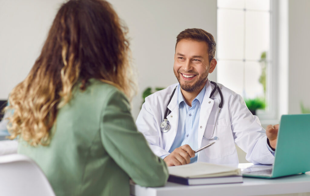 Cheerful friendly male doctor at medical office in hospital, showing examination results on laptop monitor screen and having consultation with young woman patient sitting in clinic.