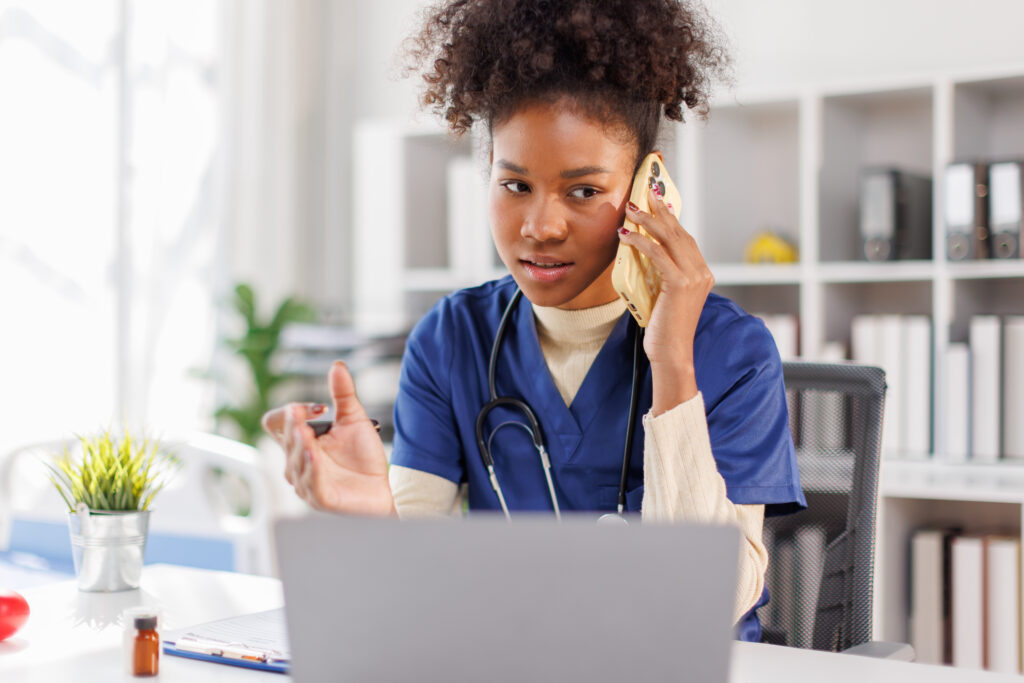 Happy friendly young woman doctor, nurse, medical worker, reception specialist in blue uniform with stethoscope sitting at desk, talking to patient on phone and checking schedule information on laptop