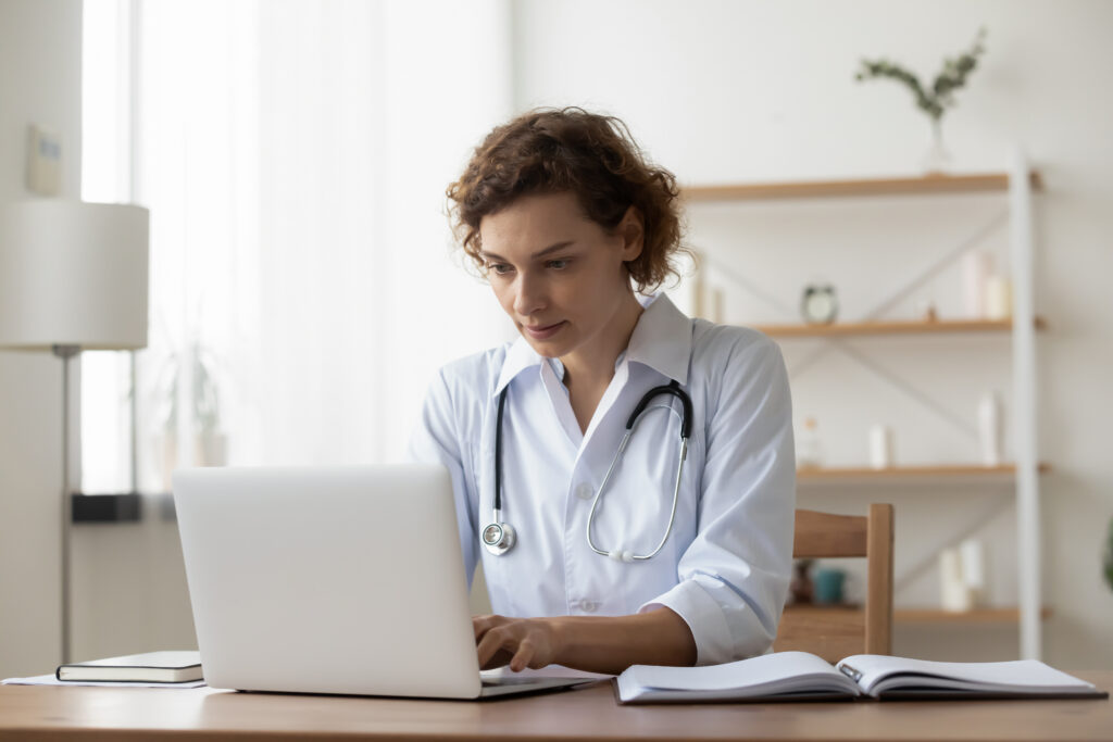 Serious woman wearing white uniform with stethoscope working on laptop, sitting at desk in hospital, focused doctor physician gp looking at computer screen, using medical apps, consulting online