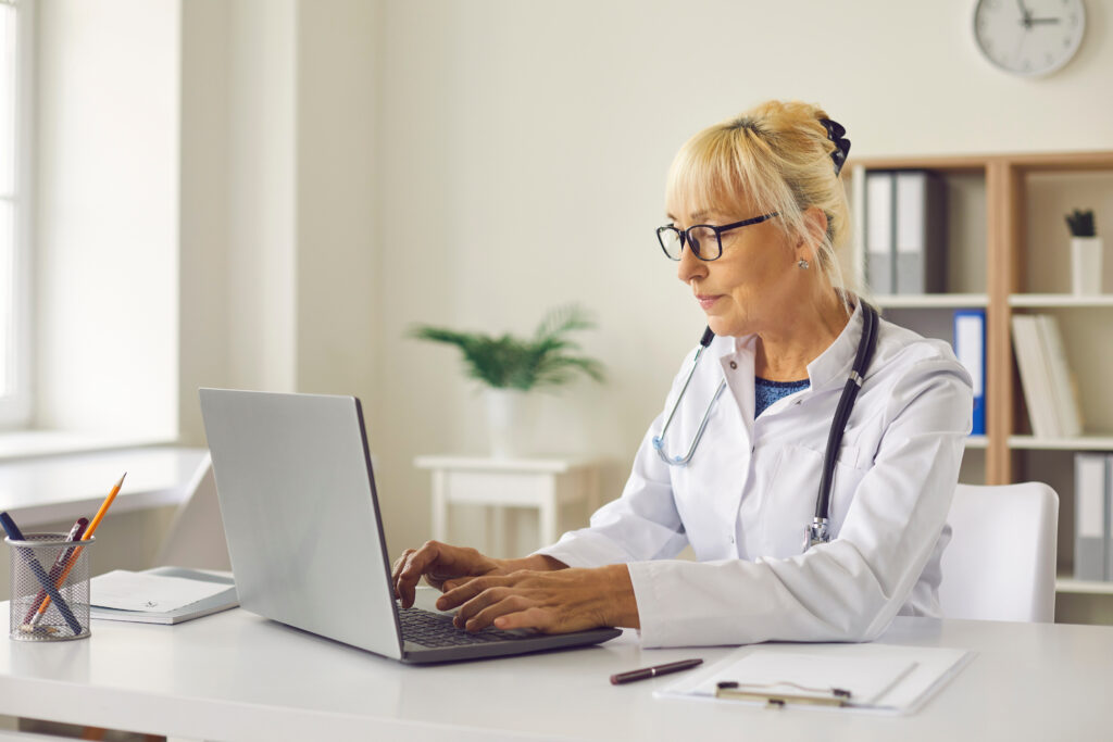 Focused senior female doctor sits in her office in front of a laptop and enters the patient's diagnosis into a medical electronic database. Concept of using modern technologies in medicine.