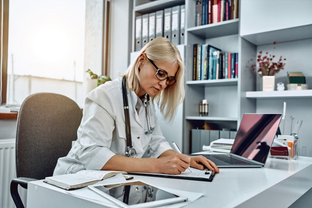 Serious female doctor using laptop and writing notes in medical journal sitting at desk. Senior woman professional medic physician wearing white coat and stethoscope working on computer at workplace