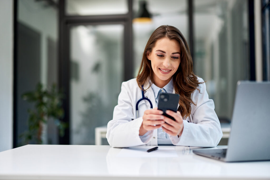 A smiling brunette female doctor texts messages on a mobile phone while sitting in front of the laptop at the office.