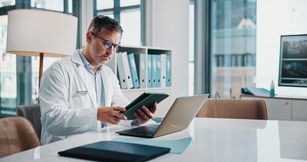 How AI Scribe Software Helps Reduce Coding Errors Healthcare, laptop and tablet with doctor man at desk in office of hospital for diagnosis, planning or research. Computer, medical and technology with mature medicine professional reading in clinic.