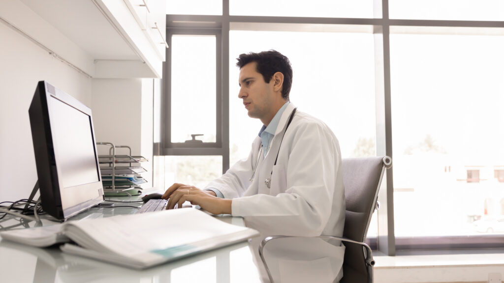 Reducing After-Hour Work With AI Clinical Documentation Focused young medical practitioner man working at computer in doctor office, typing on keyboard, writing electronic medical records, examination reports, using modern technology