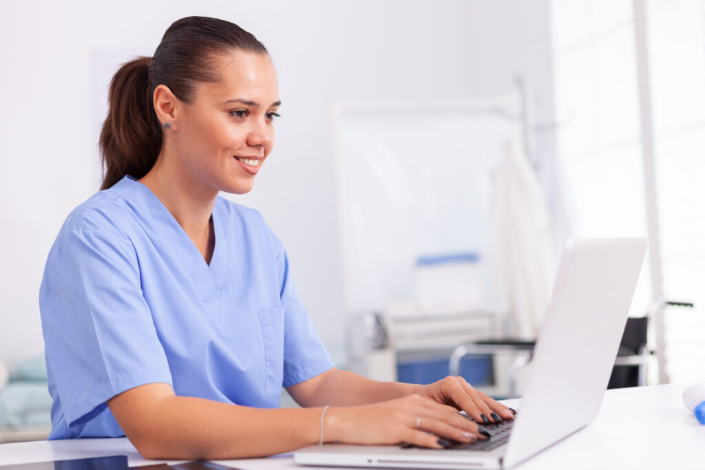 Why Revenue Cycle Intelligence Matters for Growing Practices Medical nurse in uniform using laptop sitting at desk in hospital office. Health care physician using computer in modern clinic looking at monitor, medicine, profession, scrubs.