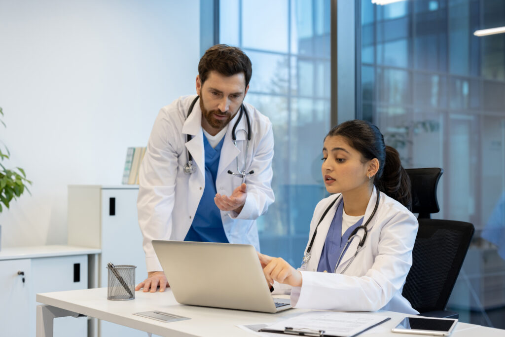 Benefits of Integrated Electronic Prior Authorization Tools Two doctors collaborate in a modern office setting, focusing on a laptop while discussing medical information and data. The scene portrays professional teamwork.