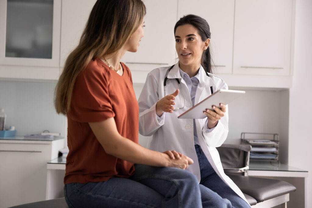 Professional Latina therapist holding modern digital tablet during conversation with woman patient, accessing electronic health record system, explain treatment plan or health test results. Healthcare
