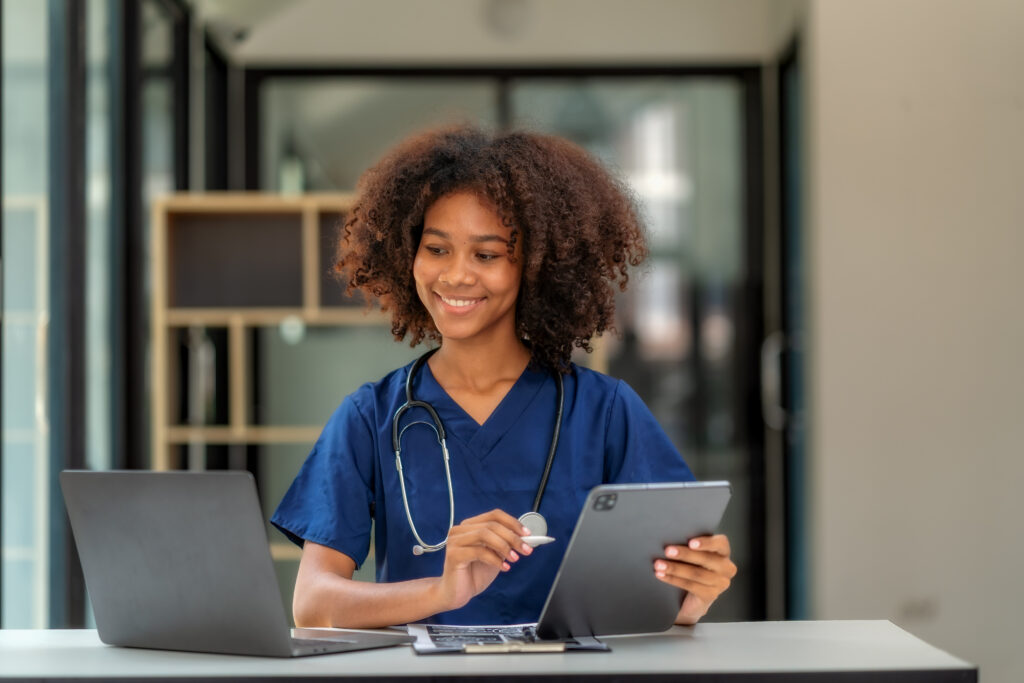 How Unified Messaging Improves Clinical Communication A cheerful young African American female doctor or nurse in blue scrubs sits at her desk, multitasking with a digital tablet, a laptop, and a medical clipboard. She is looking at the tablet with a positive smile. This image represents modern healthcare technology, telemedicine, electronic health records (EHR), and efficient medical practice in a bright, professional office setting.