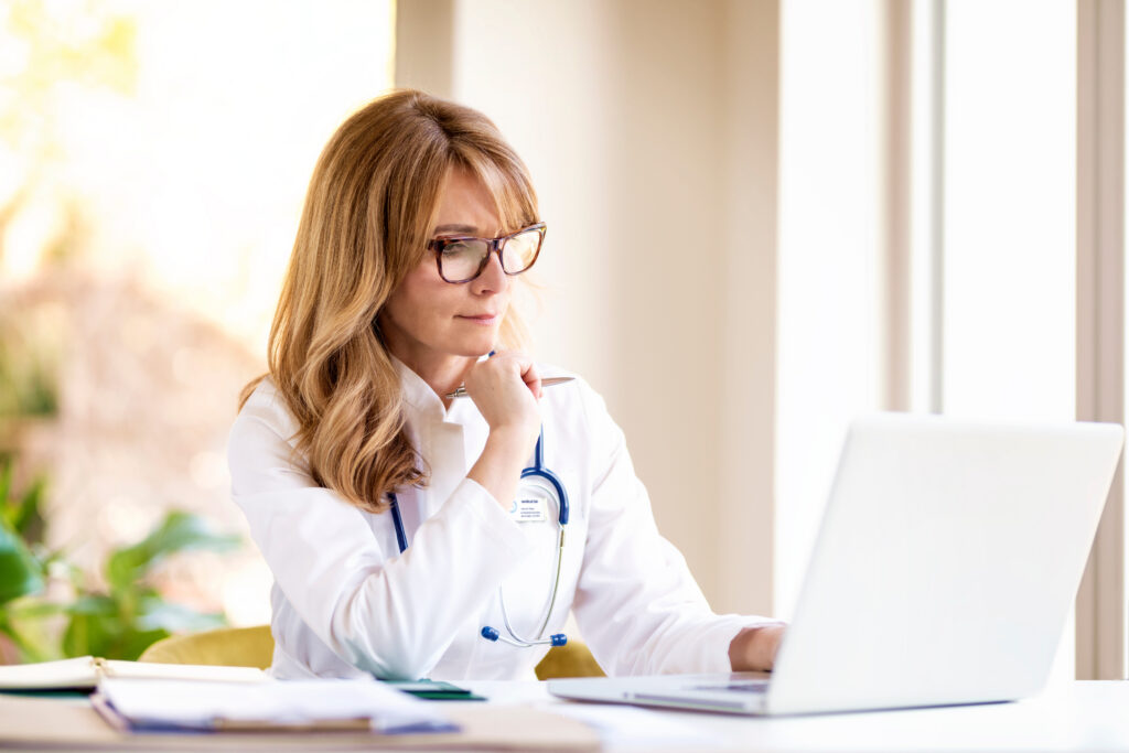 The Impact of Small Front-End Errors on the Revenue Cycle Portrait shot of thinking female doctor sitting at desk and working on laptop in doctor’s office.