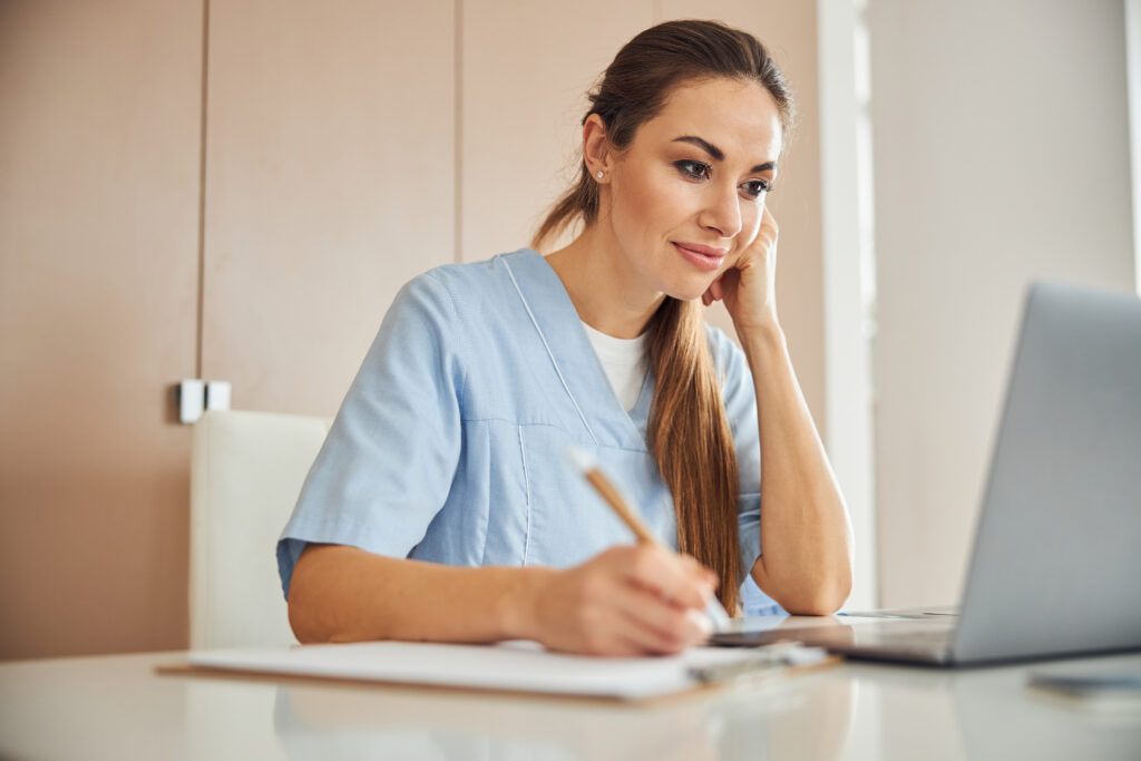 Improving Revenue Cycle Management Without Expanding Staff Charming young lady in medical scrubs sitting at the table and looking at the laptop screen