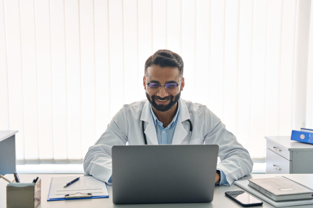 Improving Revenue Cycle Management Without Expanding Staff Indian male happy doctor medical worker in modern clinic wearing eyeglasses and white coat uniform using laptop computer having videocall chat, consulting remotely. Telemedicine health care concept.