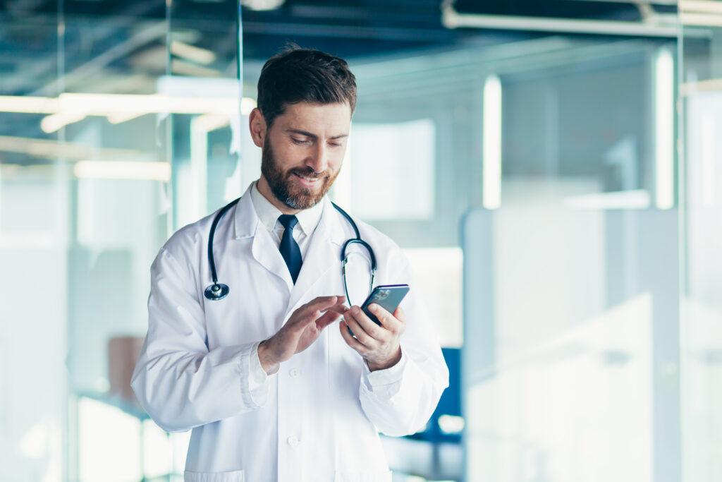 How Unified Messaging Improves Clinical Communication Male doctor in a white coat in a modern clinic reads information from a mobile phone corresponds with colleagues happy and smiles