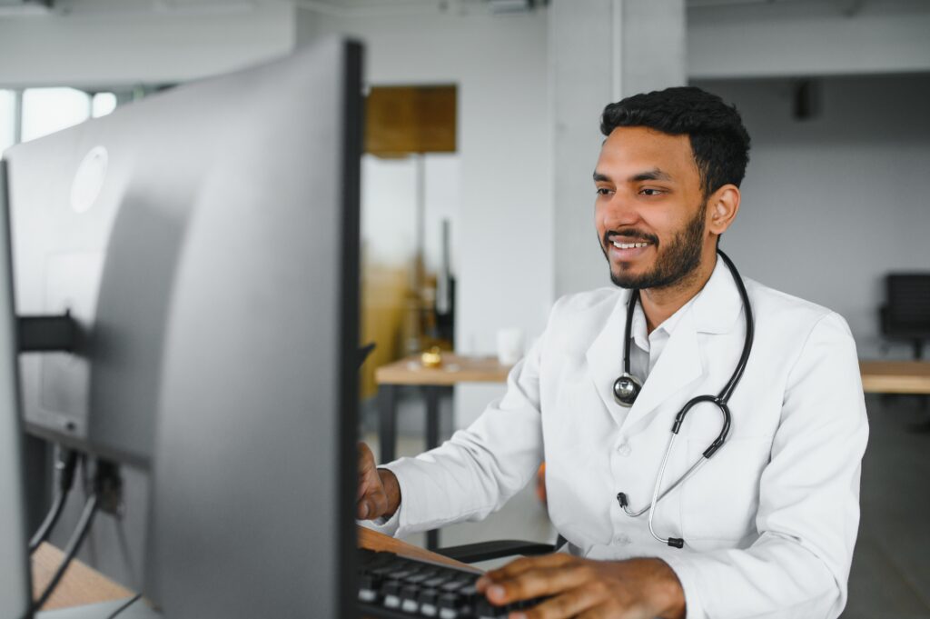 The Impact of Small Front-End Errors on the Revenue Cycle People, occupation and medicine concept. Smiling male Indian Arab doctor in white coat, sitting in medical office at desk with laptop.