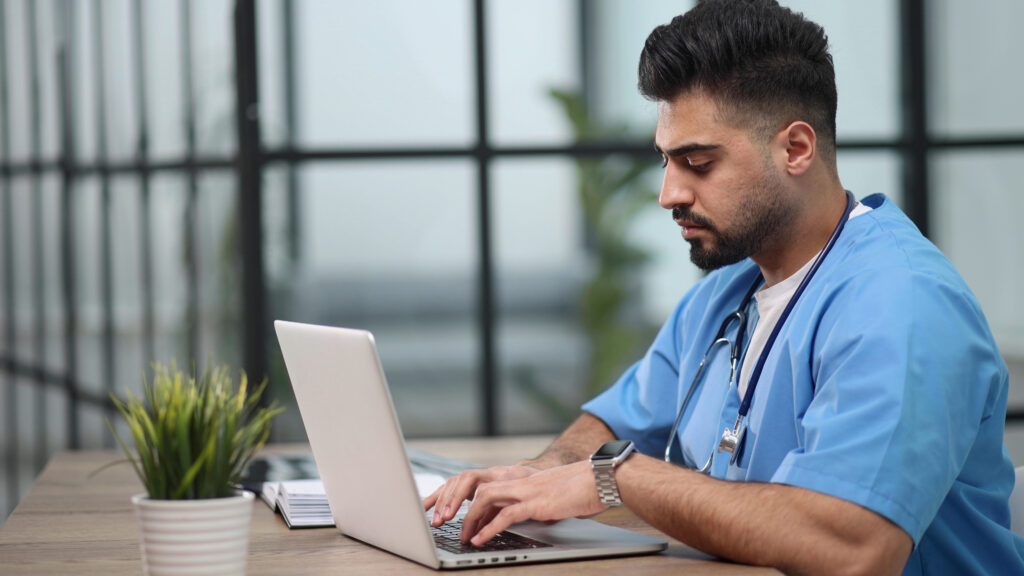Benefits of Integrated Electronic Prior Authorization Tools Young male doctor works on laptop computer at table in medical office.