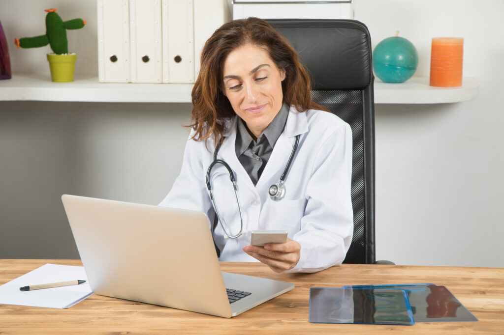 Healthcare Technology Challenges For Independent Practices brown hair doctor woman, with white gown and stethoscope, sitting in wooden table smiling, using mobile phone with portable laptop computer on desktop office