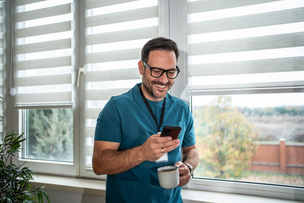 Why Your Patient Messaging Software Needs Modernization Male doctor in teal uniform smiling cheerfully while checking messages on his smartphone and drinking coffee during a break by a window in a clinic or office setting