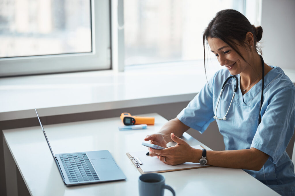 Why Your Patient Messaging Software Needs Modernization Beautiful young woman physician holding smartphone and smiling while sitting at the table with laptop and clipboard