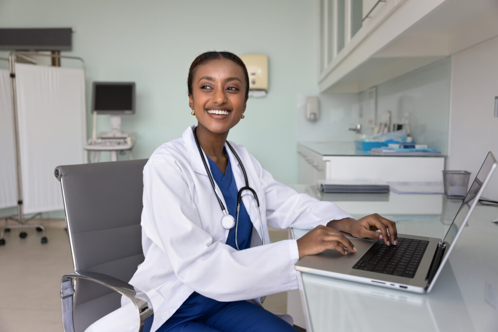 Intelligent Scheduling to Reduce No-Shows Cheerful young African American medical therapist working at laptop in hospital examination room, looking away with toothy smile, posing for portrait at clinic office workplace