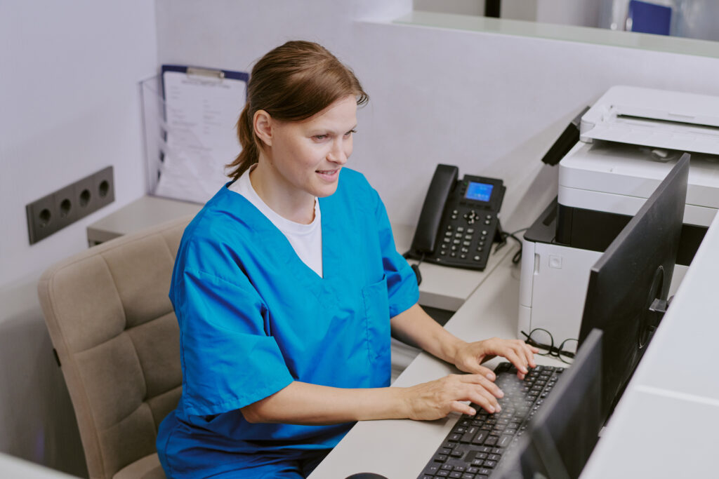 Why Front-Office Patient Communication Workflows Matter Caucasian middle aged woman wearing medical scrubs working at computer in modern office setting, sitting at desk and typing on keyboard with printer and phone visible in background